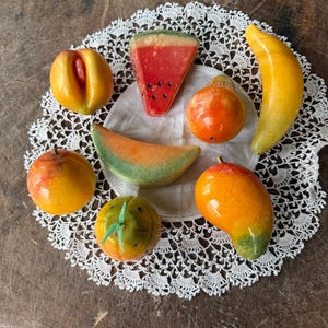 Puede incluir: Una colección de esculturas de frutas coloridas y hechas a mano, que incluyen una rodaja de sandía, un plátano, un mango y un melocotón, dispuestas sobre un mantel individual de encaje blanco. Las frutas son de material brillante y sobre fondo de madera oscura.