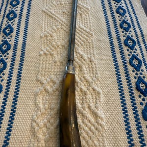 May include: A vintage knife sharpener with a dark brown horn handle and a silver metal bolster. The sharpening steel is long and cylindrical. The tool is set against a cream-colored woven background with blue accents.