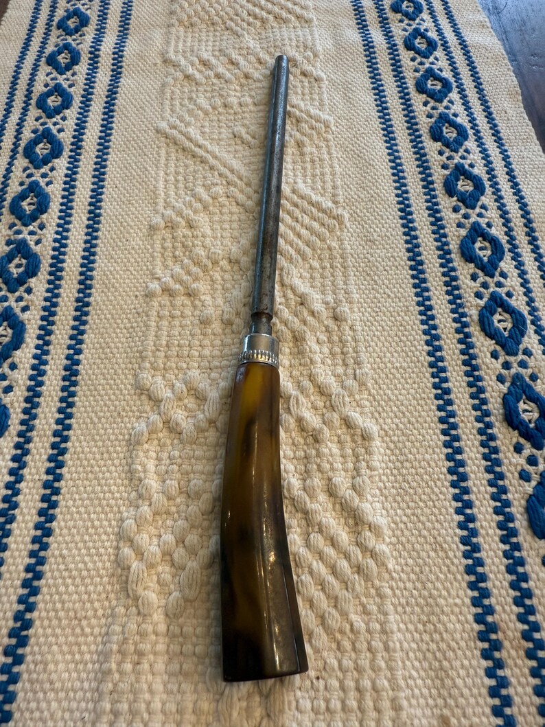 May include: A vintage knife sharpener with a dark brown horn handle and a silver metal bolster. The sharpening steel is long and cylindrical. The tool is set against a cream-colored woven background with blue accents.
