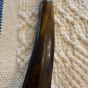 May include: Close-up of a vintage tool handle. The handle is a dark brown with yellow marbling, and a silver metal band at the top. The tool has a metal shaft extending from the top. The handle has a curved, ergonomic design.