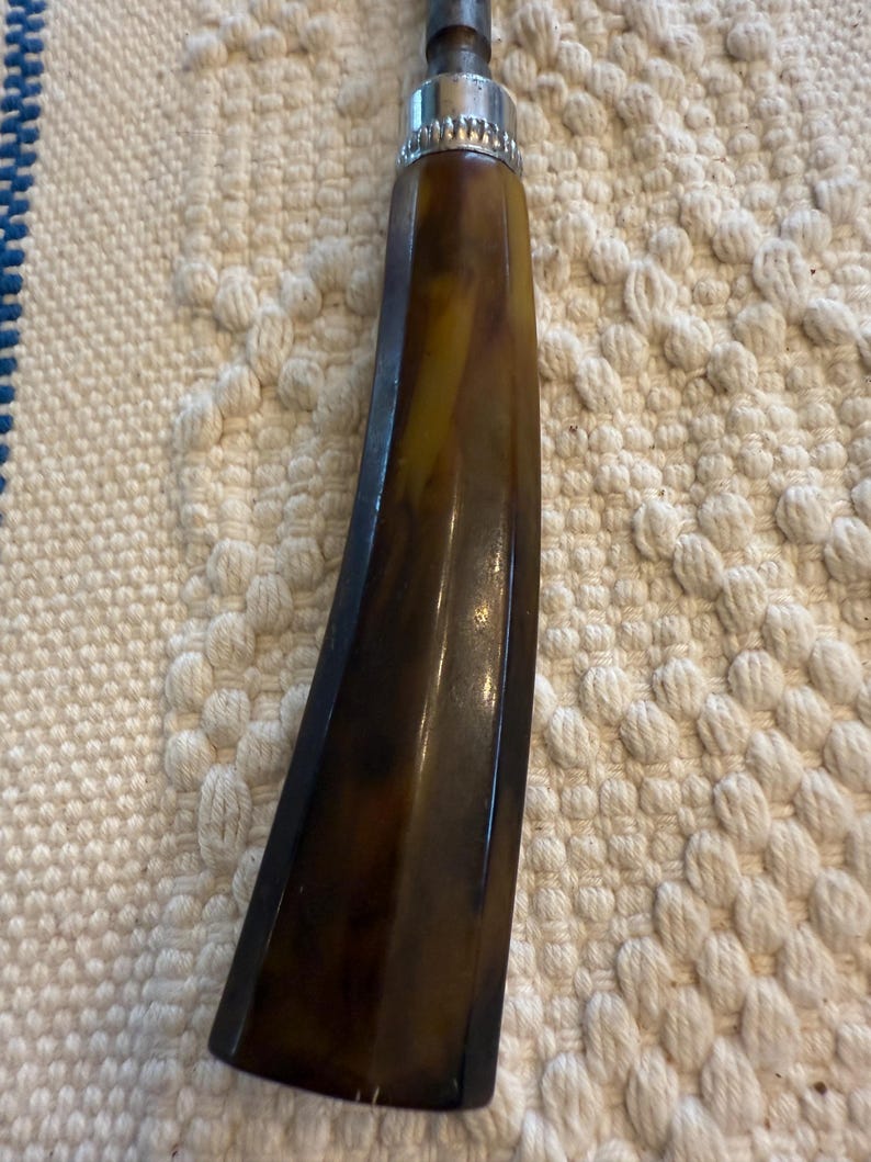 May include: Close-up of a vintage tool handle. The handle is a dark brown with yellow marbling, and a silver metal band at the top. The tool has a metal shaft extending from the top. The handle has a curved, ergonomic design.
