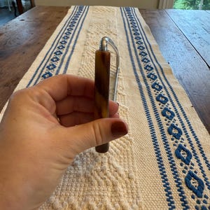 May include: A brown and silver letter opener. The handle is a marbled brown color, and the blade is silver. The letter opener is held in a hand, and the background is a white and blue patterned table runner.