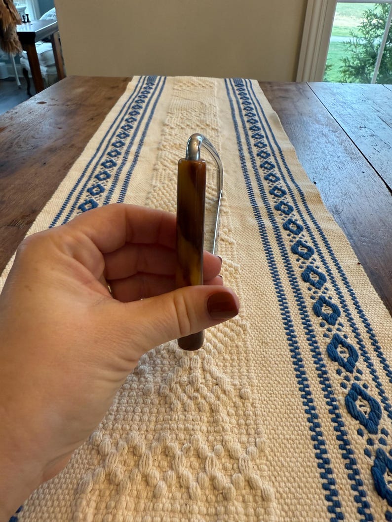 May include: A brown and silver letter opener. The handle is a marbled brown color, and the blade is silver. The letter opener is held in a hand, and the background is a white and blue patterned table runner.