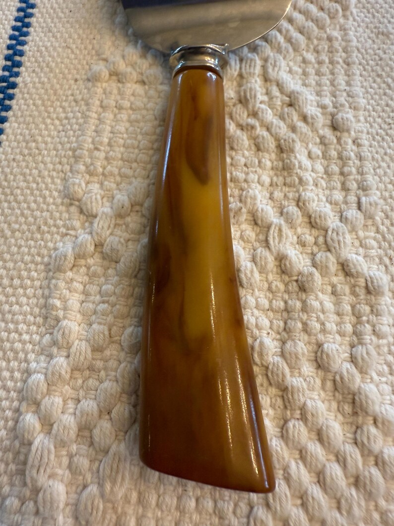 May include: Cheese knife with a stainless steel blade and a handle with a marbled amber and brown pattern. The handle has a glossy finish and a silver metal collar where it meets the blade.