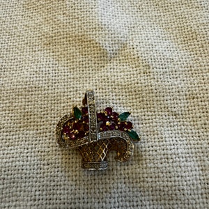 Pode incluir: Um broche dourado em forma de cesta cheia de pedras preciosas vermelhas e verdes. A cesta é decorada com um padrão de diamantes.