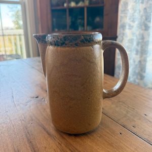 Peut inclure: Une cruche en céramique beige avec un bord et une anse marron foncé. La cruche a un bec verseur et une surface texturée. Elle est posée sur une table en bois.