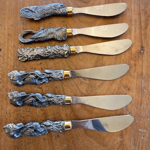 May include: A collection of six butter knives. Each knife has a silver-toned handle with a different decorative design, such as fish, elephant, and floral patterns. The blades are stainless steel with gold-coloured details.