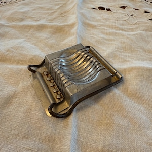 May include: A vintage egg slicer made of metal, featuring parallel wires for slicing. The slicer has a rectangular shape with a handle and is set on a beige linen surface. The metal has a worn, aged appearance.