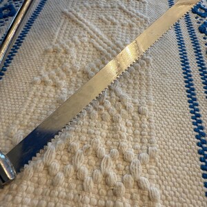 May include: A vintage metal bread knife with a serrated blade. The knife has a silver finish and a curved handle. The blade is approximately 12 inches long. The knife is resting on a white and blue patterned woven surface.