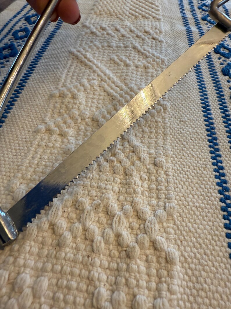 May include: A vintage metal bread knife with a serrated blade. The knife has a silver finish and a curved handle. The blade is approximately 12 inches long. The knife is resting on a white and blue patterned woven surface.