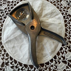 May include: A vintage metal citrus juicer with a hinged design, featuring a perforated bowl and serrated edges for efficient juice extraction. The juicer is silver-toned and rests on a white cloth doily with a delicate lace pattern.