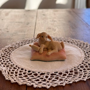 Puede incluir: Una pequeña figurita de cordero blanco acostada sobre una almohada rosa con un rodillo de madera. La figurita está sobre un mantel de ganchillo blanco sobre una mesa de madera.