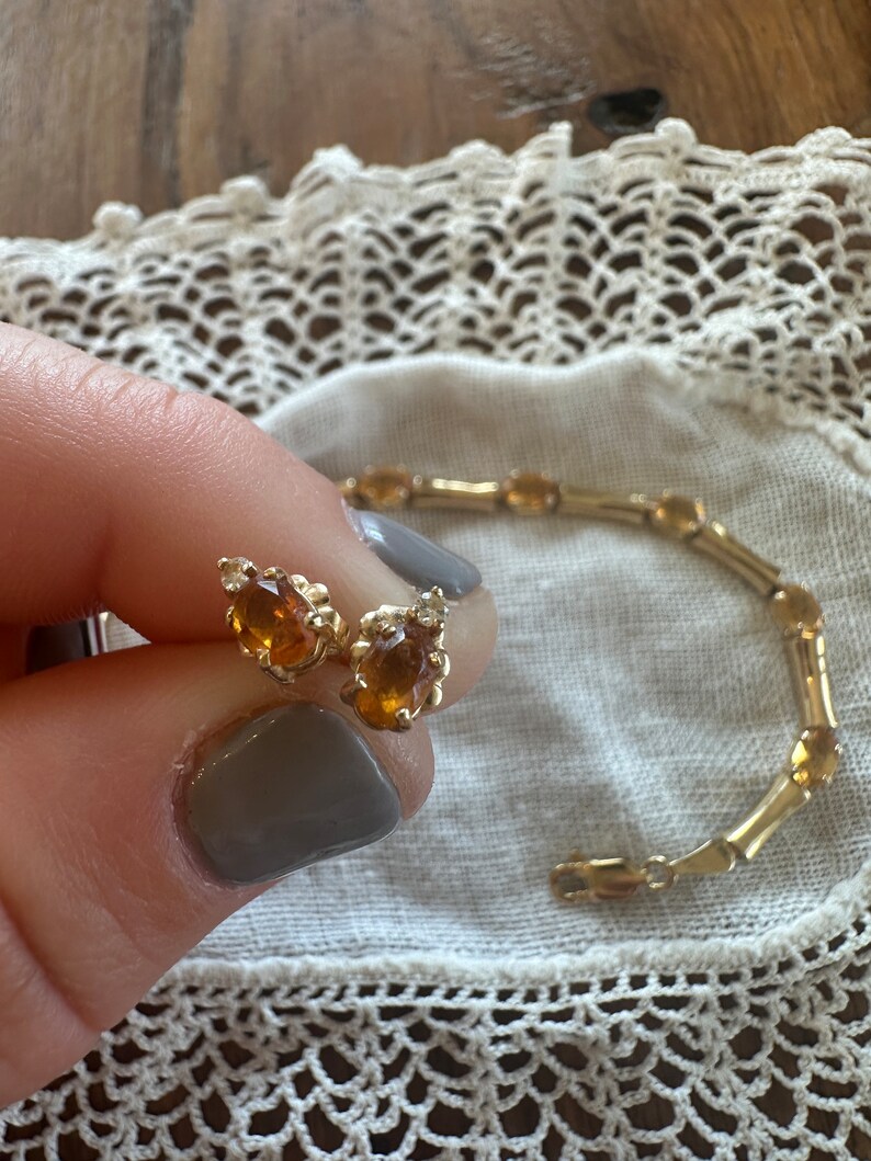 May include: A pair of gold earrings with teardrop-shaped amber stones. The earrings are shown next to a gold bracelet with oval-shaped amber stones.