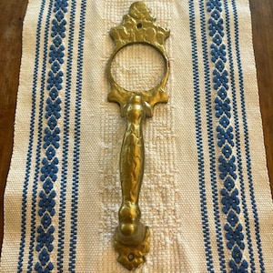 Könnte beinhalten: Ein goldfarbener Türgriff mit einem verzierten Design. Der Griff hat einen ringförmigen Griff und einen strukturierten, dekorativen Stiel.