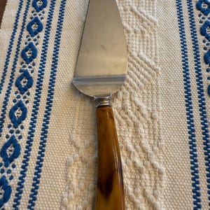 May include: A stainless steel pie server with a brown and amber-colored handle. The server has a wide, curved blade and a decorative metal band where the handle meets the blade. The server is on a cream-colored woven surface with blue embroidered accents.