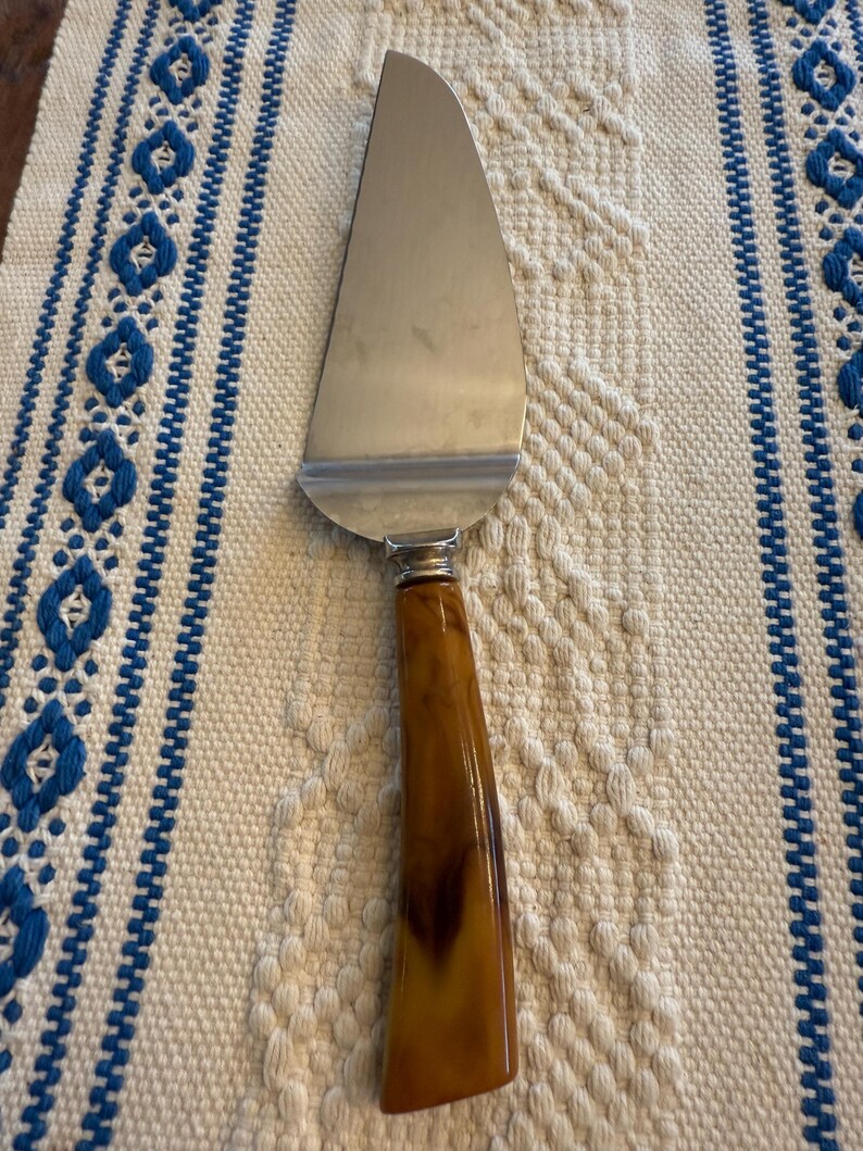 May include: A stainless steel pie server with a brown and amber-colored handle. The server has a wide, curved blade and a decorative metal band where the handle meets the blade. The server is on a cream-colored woven surface with blue embroidered accents.