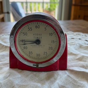 Peut inclure: Un minuteur Eastman Kodak rouge avec un cadran circulaire crème affichant des chiffres noirs de 5 à 60. L'appareil possède une lunette en argent métallique, deux aiguilles noires et le texte KODAK TIMER et EASTMAN KODAK CO. ROCHESTER, N.Y.