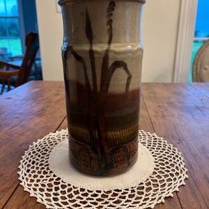May include: A brown and beige ceramic vase with a stylised plant design. The vase has a textured surface and is sitting on a white crocheted doily.