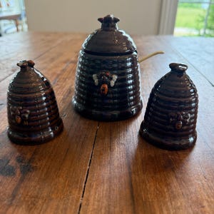 May include: A brown ceramic honey pot set with a beehive design. The set includes a lidded honey pot, a salt shaker, and a pepper shaker. Each piece features a bee emblem and a ridged texture.