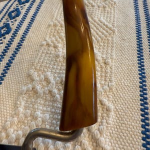 May include: Close-up of a vintage cheese slicer. The handle is a marbled amber color, and the metal frame is silver. The slicer is resting on a white and blue patterned surface.
