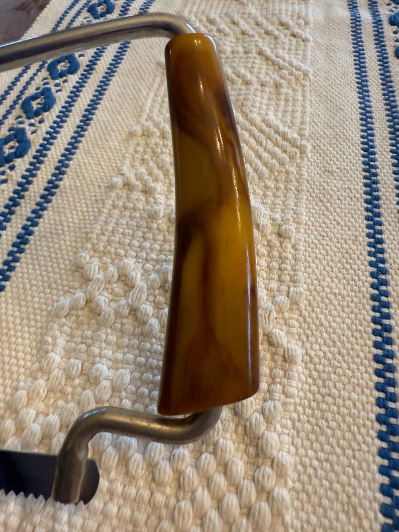 May include: Close-up of a vintage cheese slicer. The handle is a marbled amber color, and the metal frame is silver. The slicer is resting on a white and blue patterned surface.
