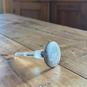 Puede incluir: Un tapón de botella de vidrio transparente con una parte superior ovalada de color plateado. La parte superior tiene un diseño grabado. El tapón está sobre una superficie de madera.