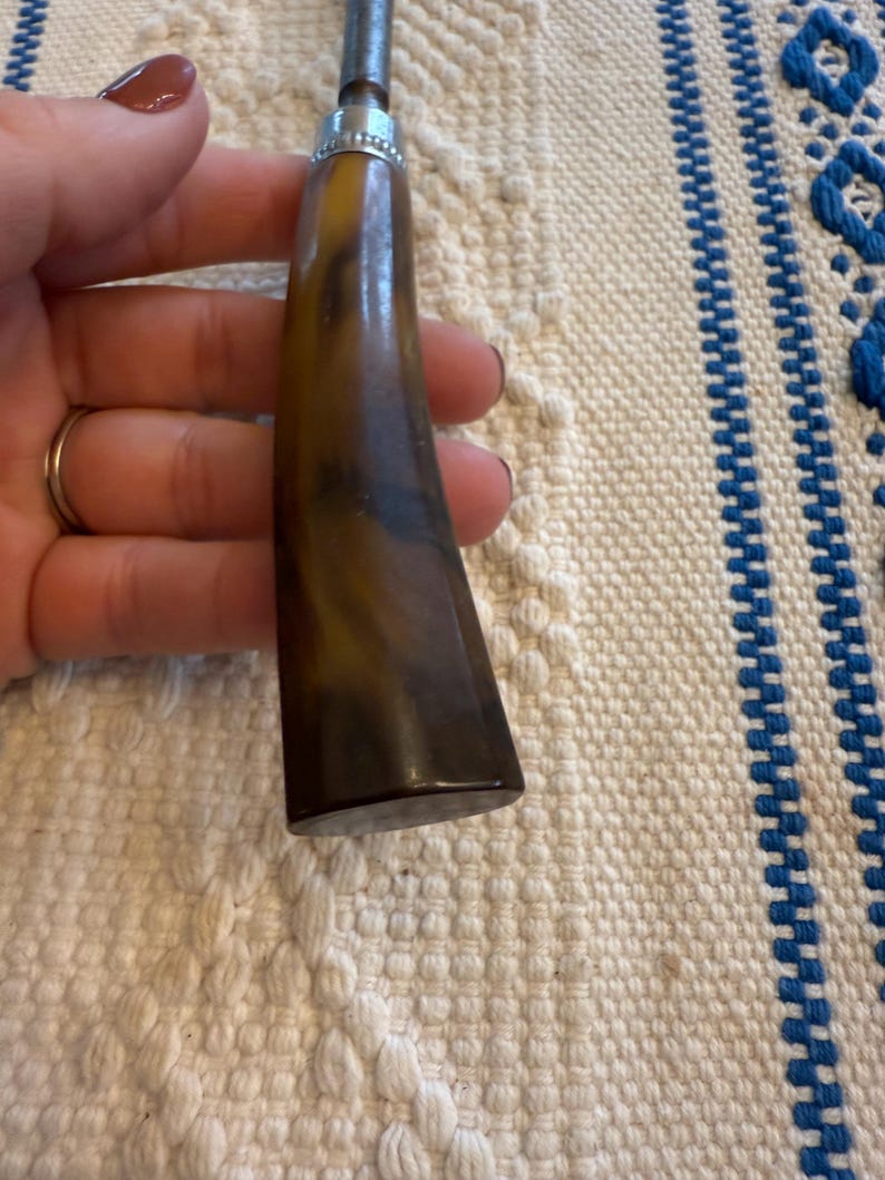 May include: A vintage tool with a dark brown, marbled handle and a silver metal top. The handle has a curved shape and a polished finish. The tool's metal portion is a dull silver color. The background is a cream-colored fabric with blue stripes.