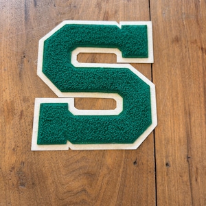 May include: A varsity-style letter S patch featuring a textured, dark green chenille center with a thick, smooth white felt border. The letter has a block-style font with sharp, angular edges and a raised, fuzzy surface texture on the green portion.