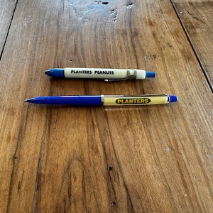 以下が含まれることがあります： プランターピーナッツと印刷された、青と白のペンが2本。ペンは木製の表面の上に置かれています。