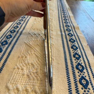May include: A stainless steel kitchen utensil, possibly tongs, with a wooden handle. The utensil is held up against a white and blue patterned tablecloth.