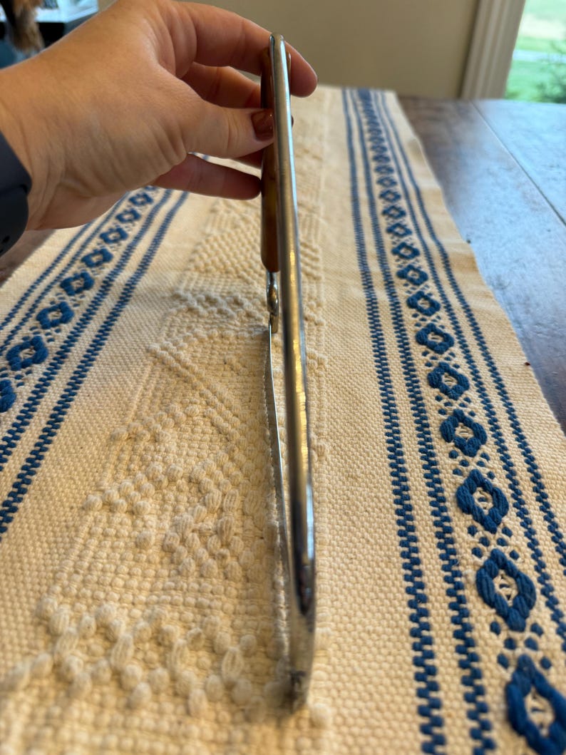 May include: A stainless steel kitchen utensil, possibly tongs, with a wooden handle. The utensil is held up against a white and blue patterned tablecloth.