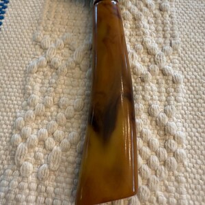 May include: Cheese slicer with a stainless steel blade and a handle made of brown and amber-colored marbled material. The handle has a rounded, ergonomic shape.