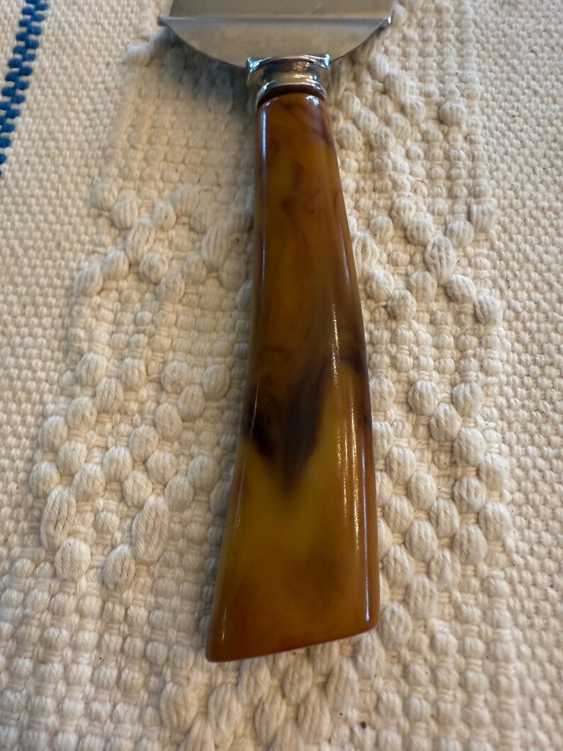 May include: Cheese slicer with a stainless steel blade and a handle made of brown and amber-colored marbled material. The handle has a rounded, ergonomic shape.
