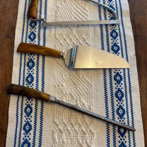May include: A collection of vintage serving utensils with brown handles and stainless steel blades. Includes a cake server, a carving saw, and a long, thin serving tool. The items are arranged on a white and blue patterned tablecloth.
