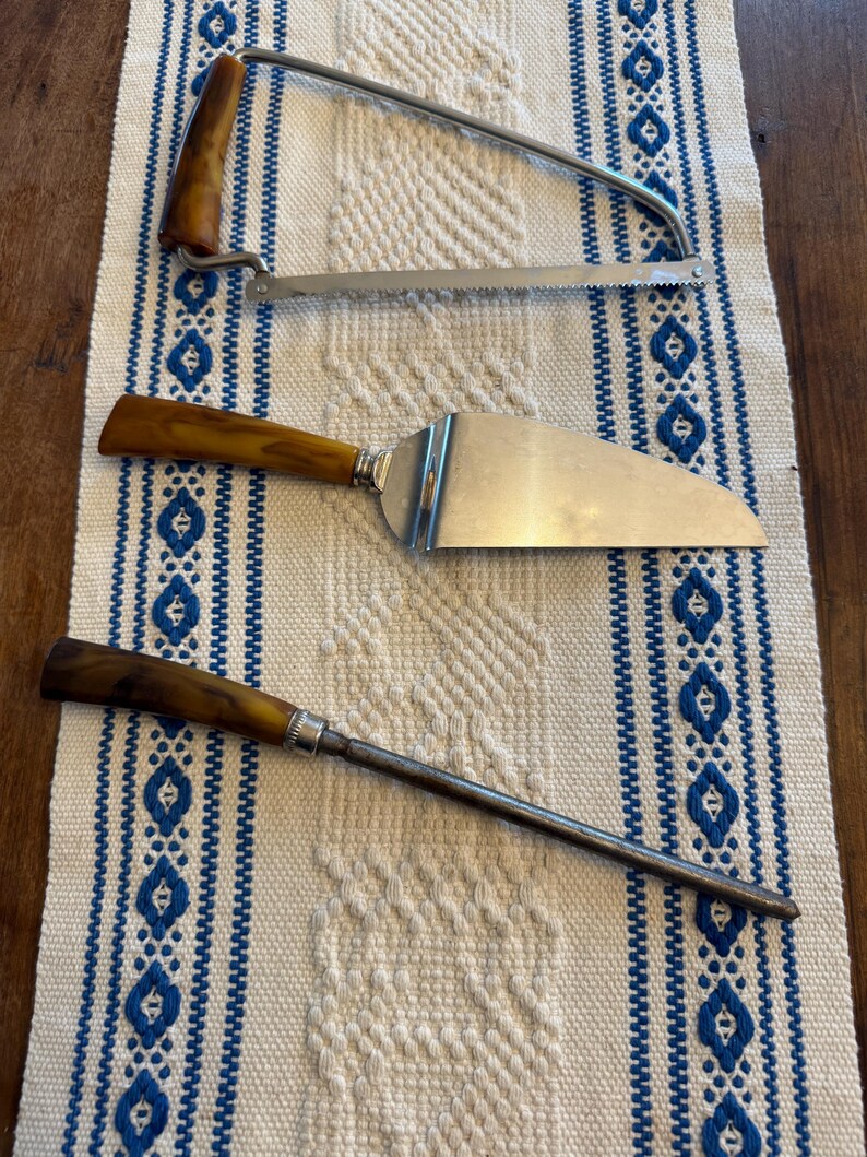 May include: A collection of vintage serving utensils with brown handles and stainless steel blades. Includes a cake server, a carving saw, and a long, thin serving tool. The items are arranged on a white and blue patterned tablecloth.