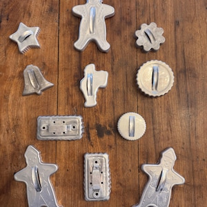 Pode incluir: Uma coleção de 12 cortadores de biscoitos de metal vintage em várias formas, incluindo figuras de pão de gengibre, estrelas, sinos e um círculo. Os cortadores são prateados e têm um aspecto rústico.