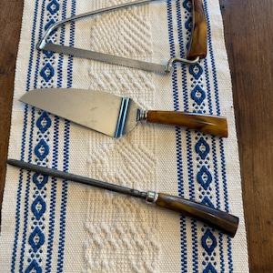 May include: Three vintage serving utensils with brown handles and stainless steel blades. Includes a cake server, a carving saw, and a sharpening steel. The items are arranged on a white and blue patterned table runner.