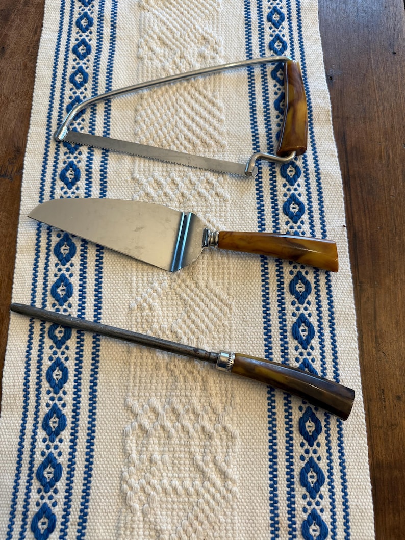 May include: Three vintage serving utensils with brown handles and stainless steel blades. Includes a cake server, a carving saw, and a sharpening steel. The items are arranged on a white and blue patterned table runner.
