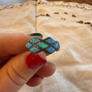 Puede incluir: Anillo de plata con una serie de piedras azules similares al ópalo engastadas en un patrón geométrico. Las piedras tienen una calidad brillante e iridiscente, que refleja tonos de azul y verde. El diseño del anillo es una serie de formas de diamante conectadas.