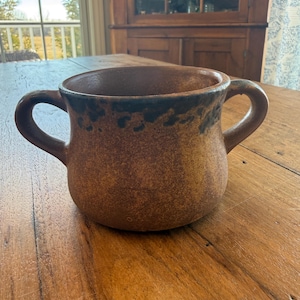 Peut inclure: Une tasse en céramique marron avec deux anses, présentant une surface texturée et un bord marron foncé. La tasse est posée sur une table en bois. Cette tasse artisanale est adaptée aux boissons chaudes comme le thé ou le café, et a un aspect rustique.