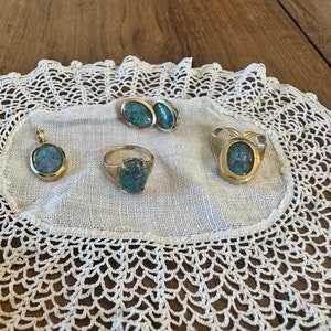 Puede incluir: Un conjunto de cuatro piezas de joyería de oro con piedras de ópalo. El conjunto incluye un colgante, un anillo y un par de pendientes. La joyería se muestra en un encaje de ganchillo blanco.