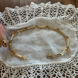 May include: A gold bracelet with a chain link design and amber-colored stones. The bracelet is on a white crocheted doily.