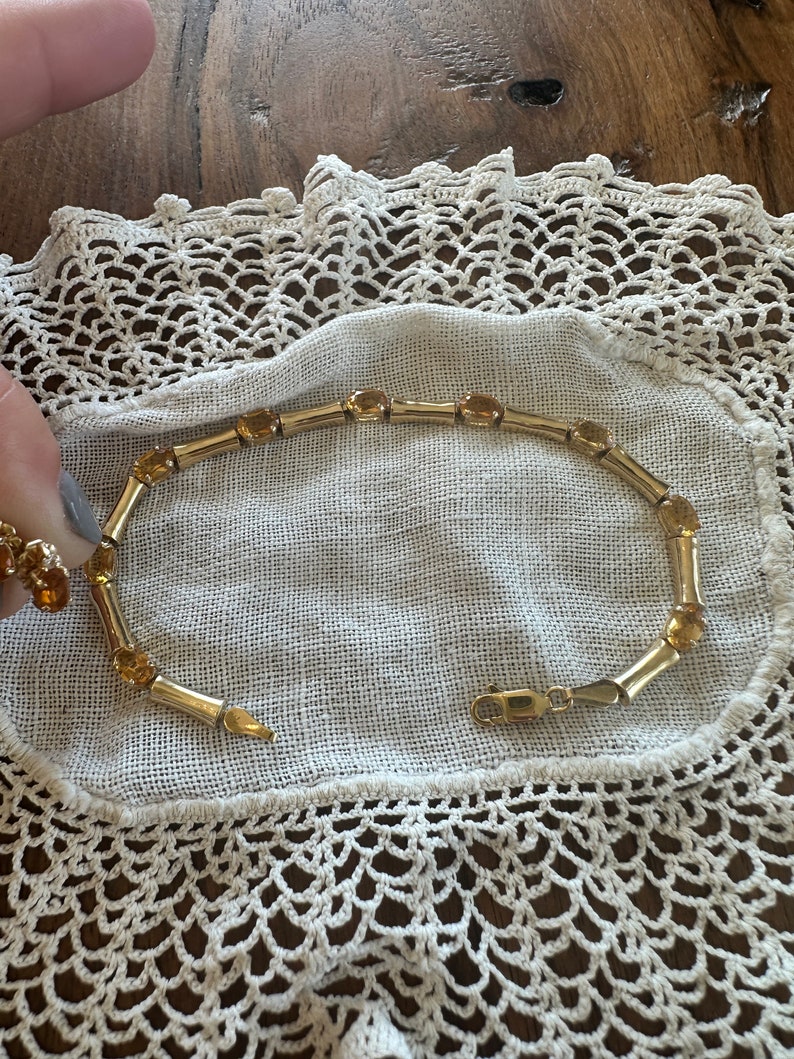 May include: A gold bracelet with a chain link design and amber-colored stones. The bracelet is on a white crocheted doily.