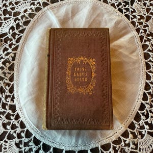 Può includere: Un libro vintage marrone con lettere dorate sulla copertina. Il titolo del libro è "Young Lady's Guide".