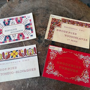 Op de afbeelding: Vier vintage borduurpatroonboeken met kleurrijke bloemen en geometrische ontwerpen. De boeken hebben titels in verschillende talen, waaronder "Bordados Bulgaros" en "Broderies Yougoslaves."
