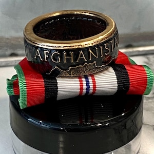 May include: A black ring with gold lettering that reads "AFGHANISTAN" on a black and green ribbon with a white ribbon with red and blue stripes on top. The ring is sitting on a black and clear container.