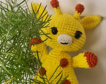 May include: A handmade yellow crochet giraffe with brown spots and accents. The giraffe has black eyes, a white muzzle, and brown-tipped ears. The giraffe is partially obscured by green foliage.