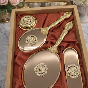 May include: A vanity set in a wooden box, featuring a round compact, two hand mirrors, and a rectangular tray. The set is gold-toned with ornate handles and floral crystal accents. The items rest on a rust-colored fabric.