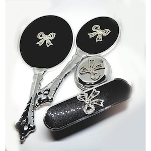 May include: A collection of vanity items: two hand mirrors, a compact, and a case. Each has a black surface with a silver bow. The handles and compact are silver. The case is black with glitter.