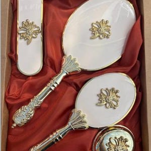 Könnte beinhalten: Ein weiß-goldenes Kosmetikset mit zwei ovalen Handspiegeln, einer rechteckigen Bürste und einem runden Puderetui. Jedes Stück weist ein goldenes Blumenmuster auf. Das Set wird in einer Box mit rotem Satinfutter präsentiert.